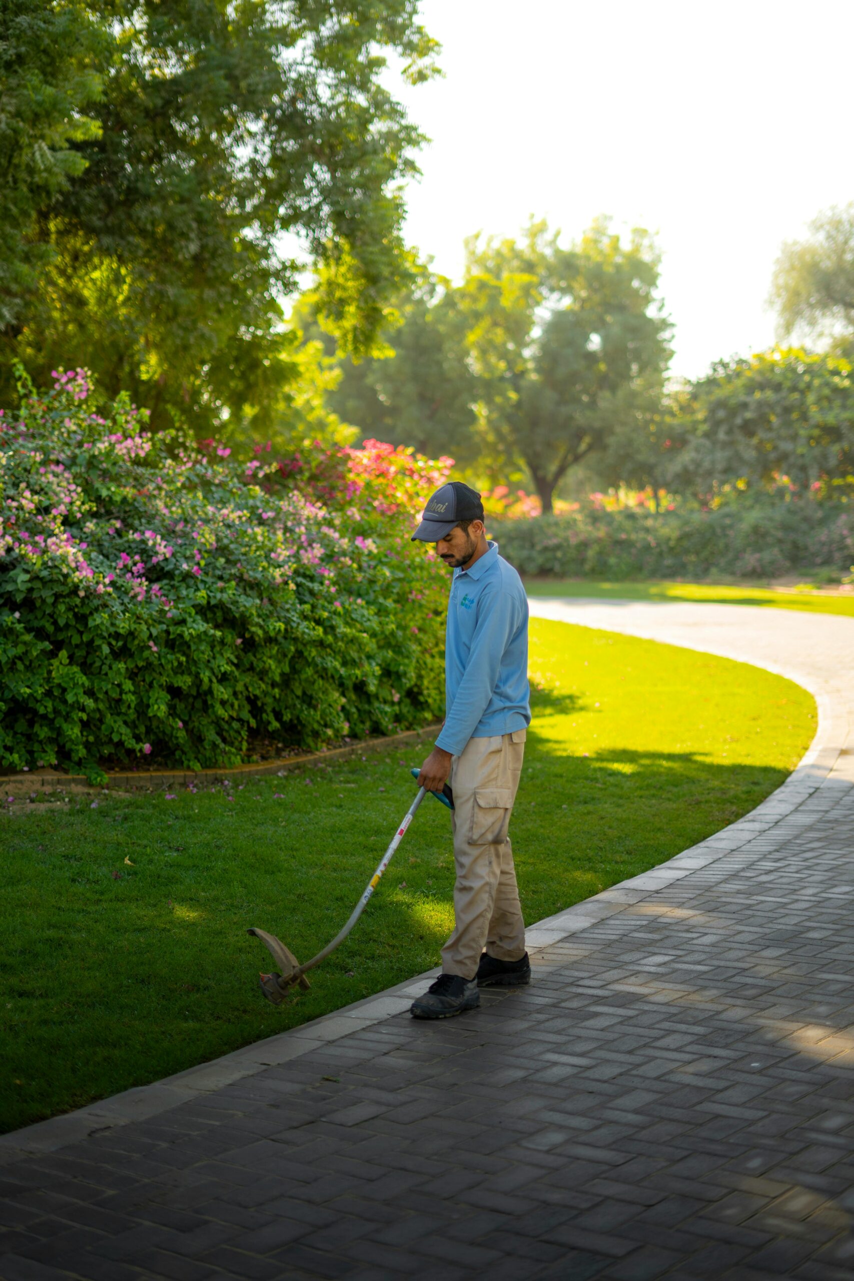 Man trimming lawn in a sunny Dubai park, surrounded by lush greenery and flowers.